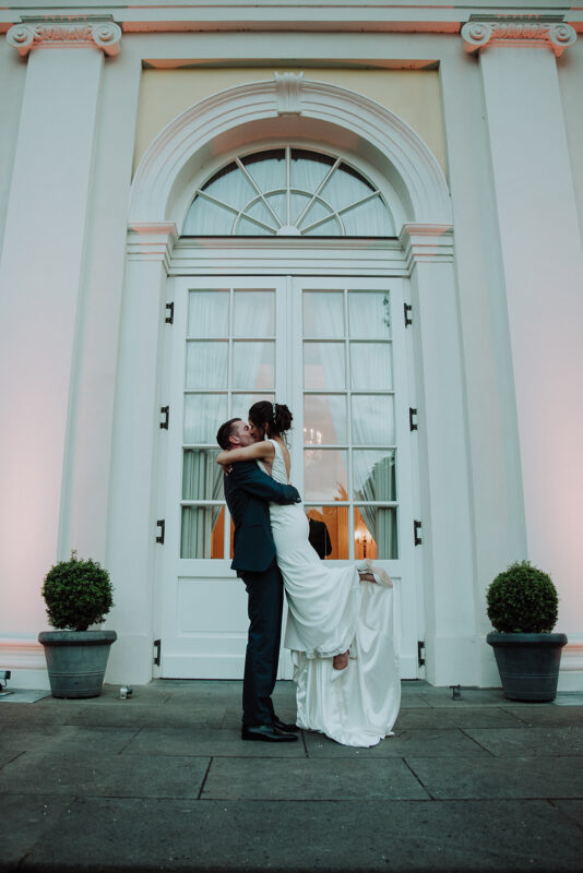 Marc und Jasmin küssen sich auf der Terrasse der Redoute, er habt sie im weißen Hochzeitskleid dabei noch.