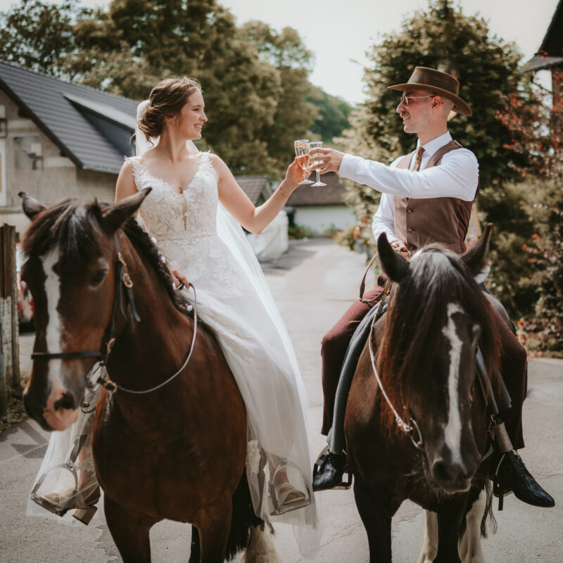 Ricci sitzt im Brautkleid auf einem braunen Pferd. Willi mit weißem Hemd, Krawatte, Weste und breitkrempigem Hut auf einem zweiten. Jeder hat ein Sektglas in der Hand und sie stoßen an.
